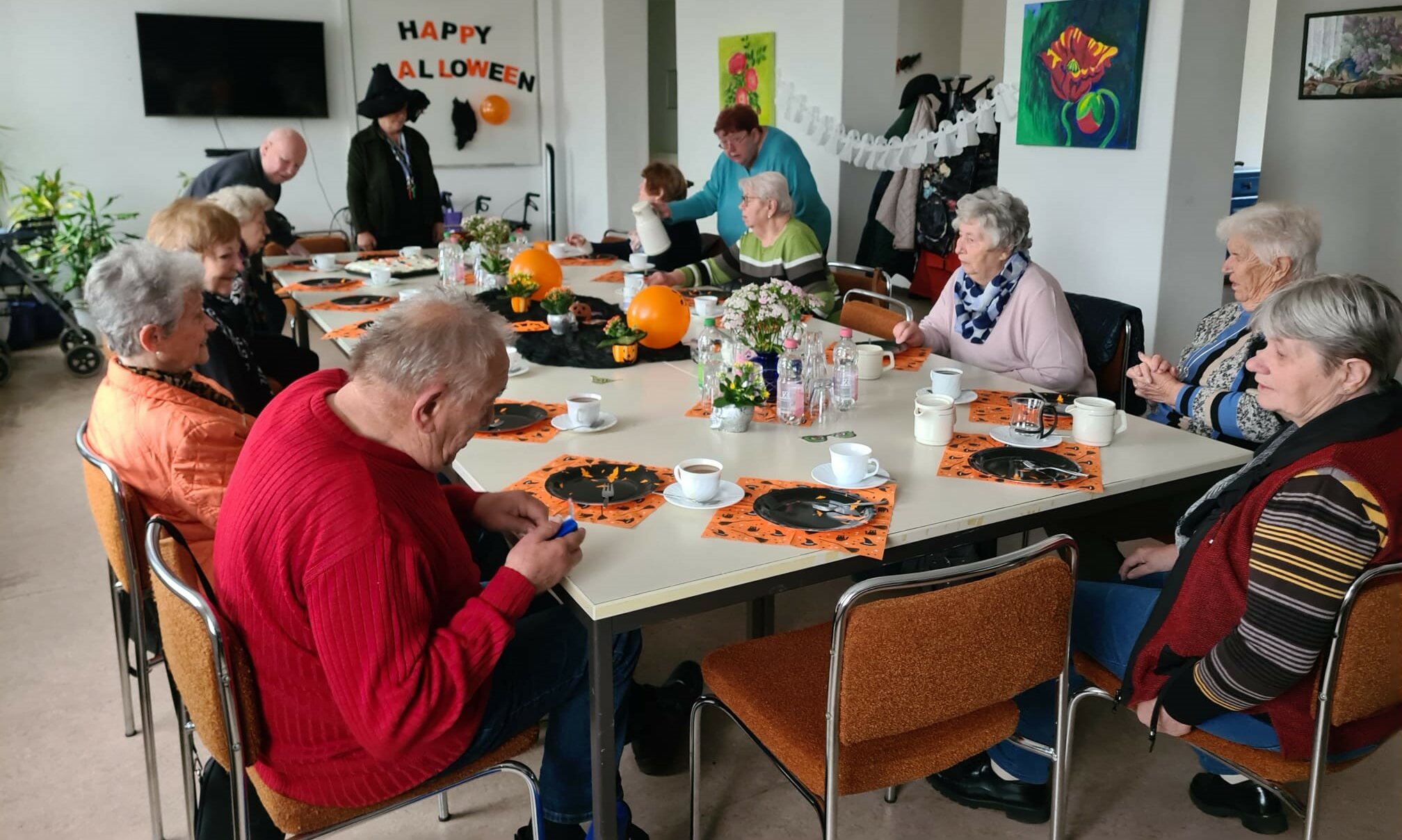 Eine Gruppe älterer Menschen sitzt an einem Tisch. Der Raum ist für Helloween dekoriert.  