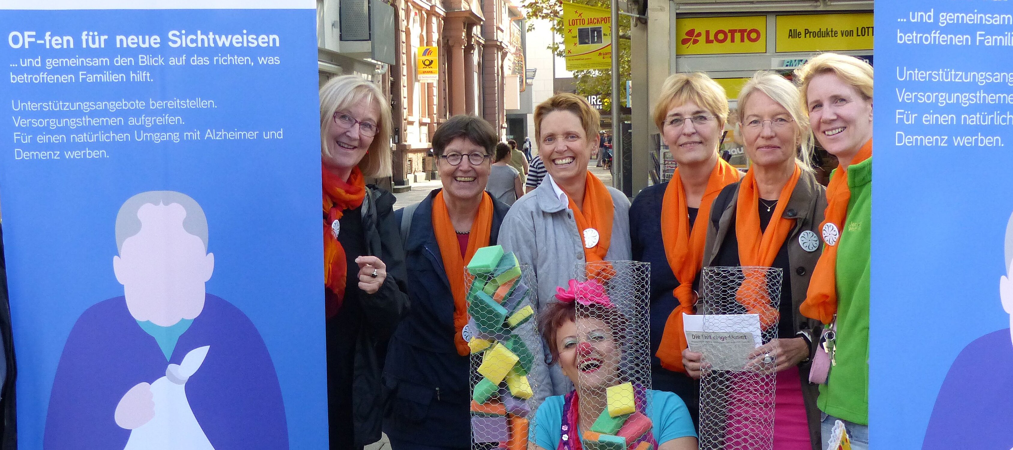 Eine Gruppe Frauen mit orangenen Schals und Aktionsmaterialien zwischen zwei Plakaten des Demenznetzwerkes Offenbach.