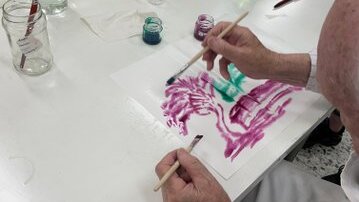 Foto: An einem Tisch malt eine ältere Person mit Aquarellfarben