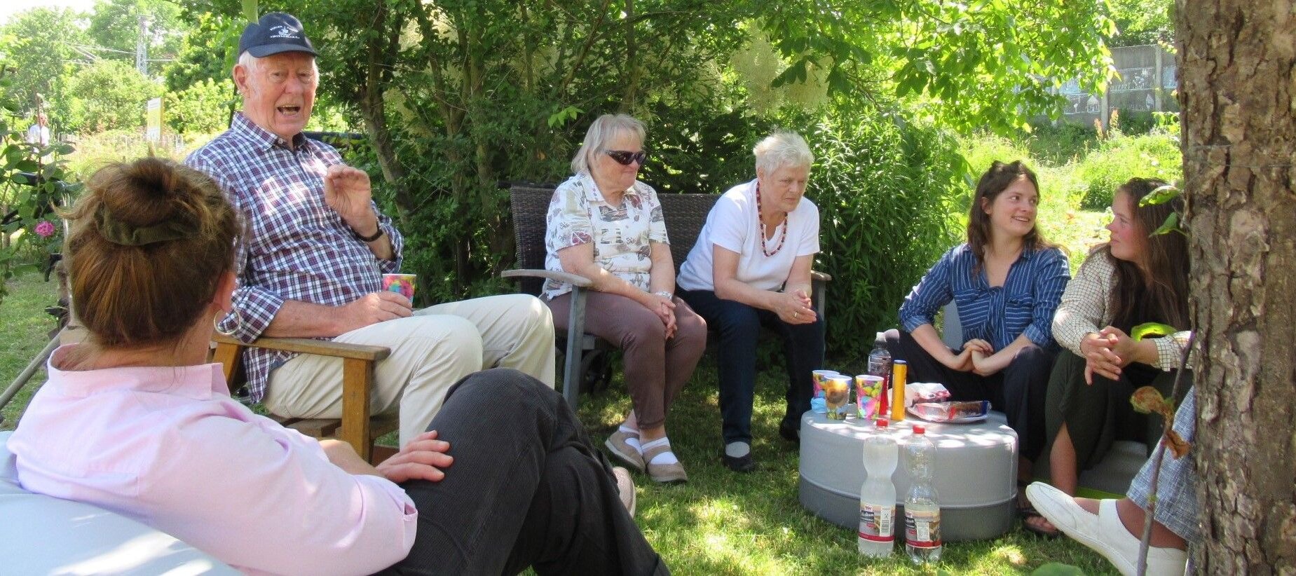 Foto: Sechs Personen sitzen in einem Halbkreis im Garten unter einem Baum um einen kleinen Hocker mit Bechern und Getränken