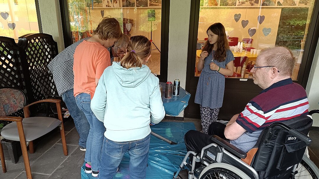 Foto: 3 Mädchen und ein Senior im Rollstuhl schauen einer älteren Dame bei einer Tätigkeit an einem mit blauer Folie abgeseckten Tisch zu.
