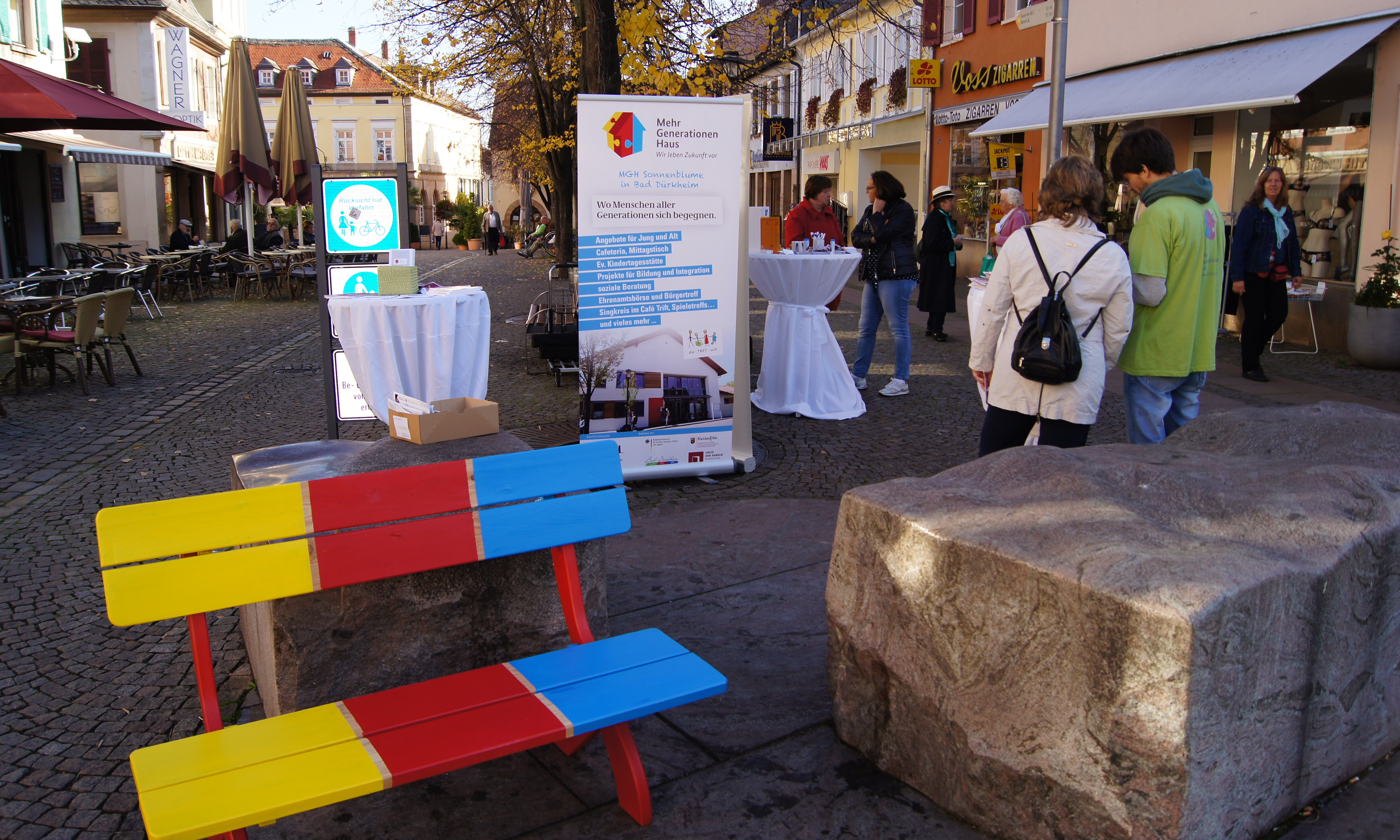 Eine draußen stehende bunte Sitzbank. Daneben ist ein größerer Steinblock. Im Hintergrund sind Personen, Stehtische und ein Roll Up vom Mehrgenerationenhaus.