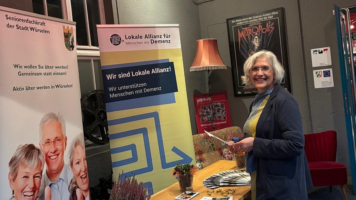 Foto: Frau steht neben Holztisch mit Broschüren in einem Kinovorraum und hält einen Flyer in der Hand. Hinter dem Holztisch stehen 2 Rollups.