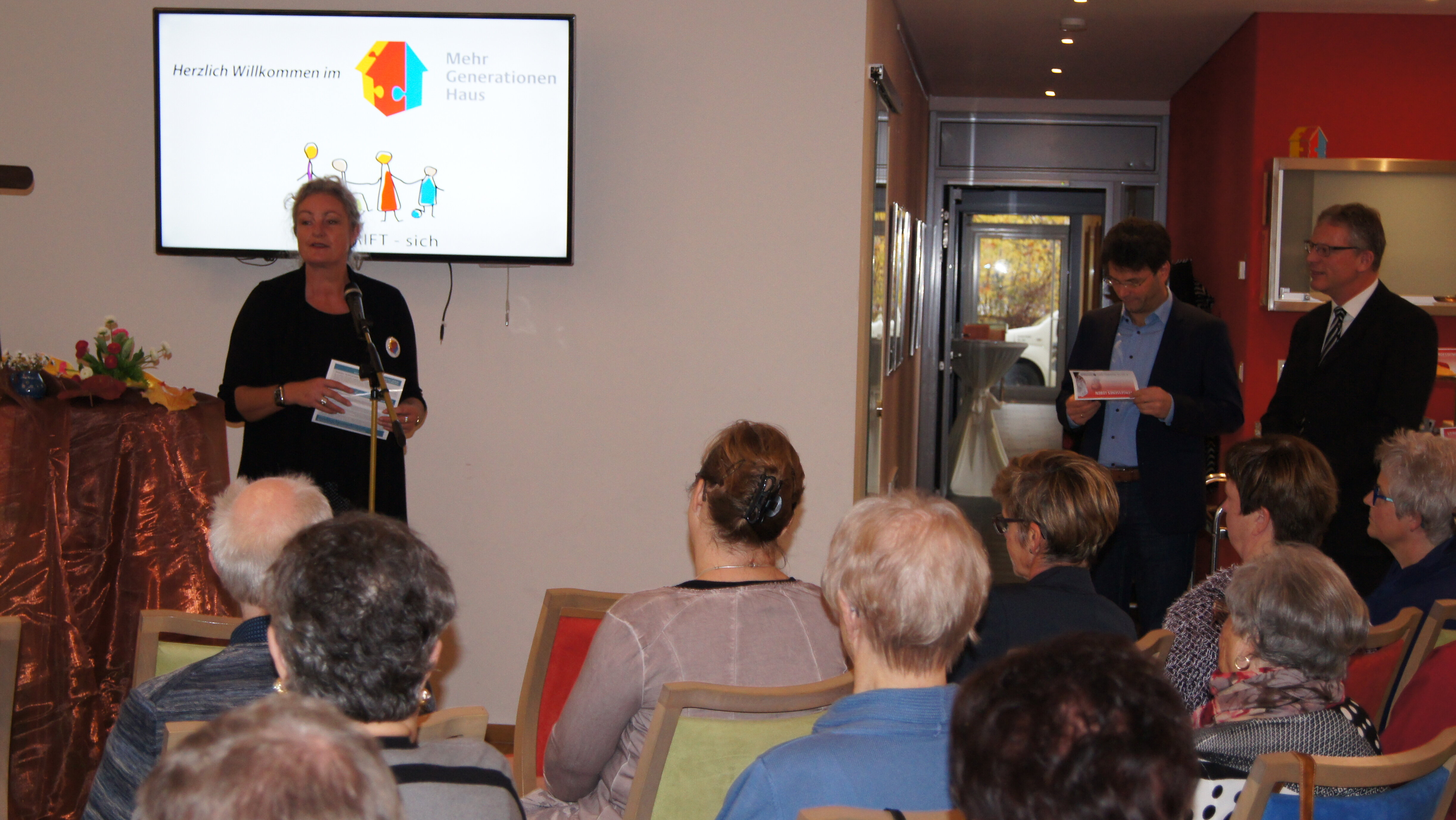 Foto: Personen, die in Stuhlreihen sitzen. Vorne steht eine Frau und hält ein Grußwort.