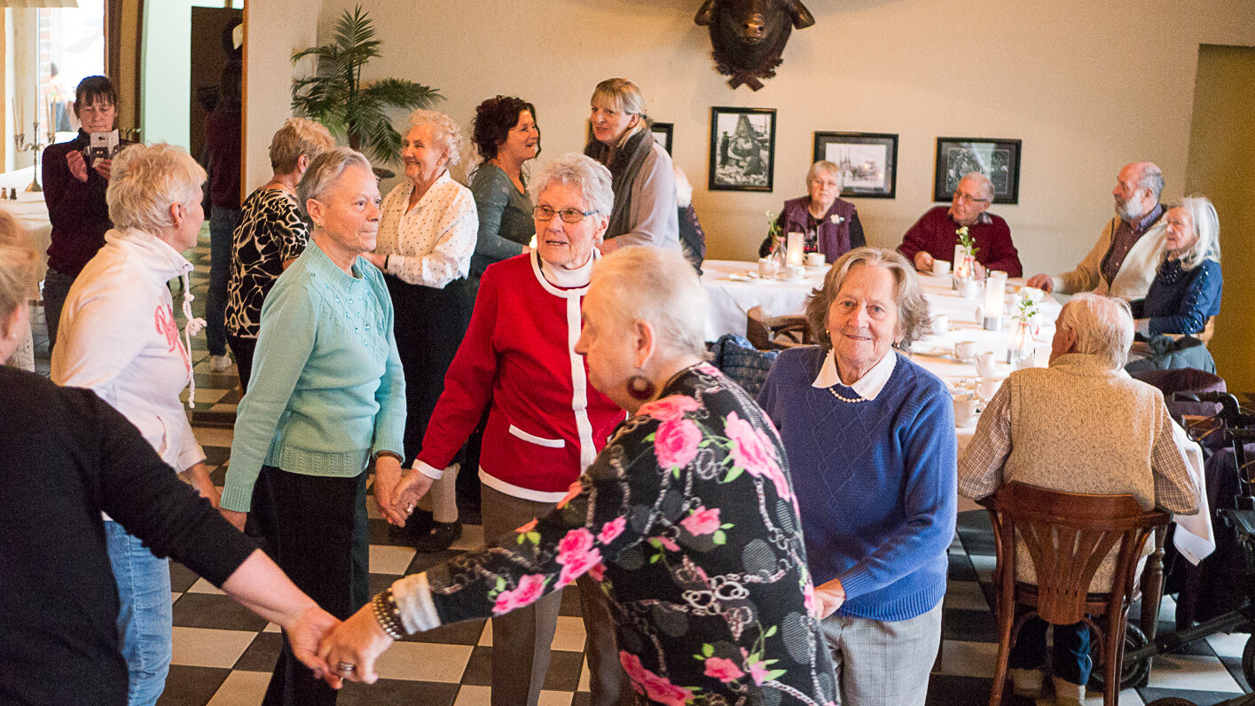 Foto: Sechs ältere Damen halten sich an den Händen und drehen sich im Kreis. Im Hintergrund sieht man weitere Personen, die zuschauen oder an einem Tisch sitzen.  