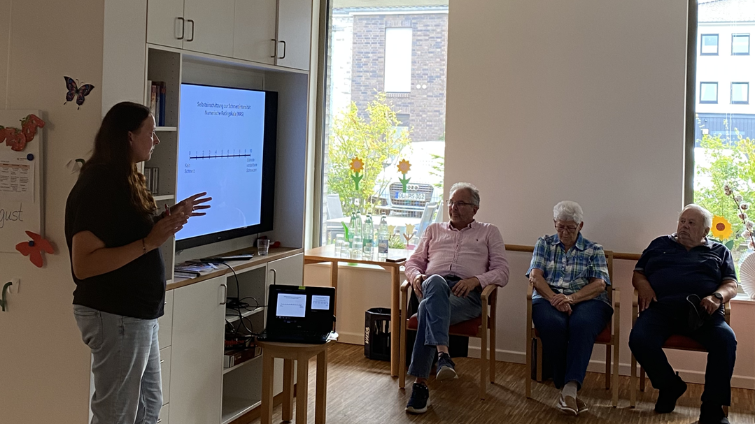 Foto: Eine Frau hält in einem Seminarraum einen Vortrag, in einem Schrank hinter ihr ein großer Bildschirm mit Ihrer Online-Präsentation, vor ihr sitzen drei Zuhörer.