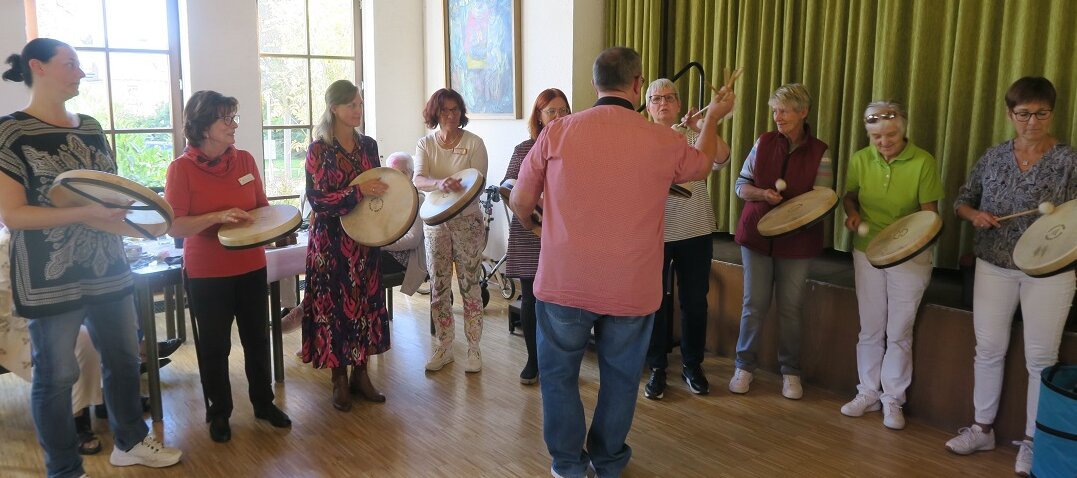 Menschen im Kreis, die mit flachen, kreisförmigen Trommeln musizieren. Vor ihnen steht ein Dirigent. 