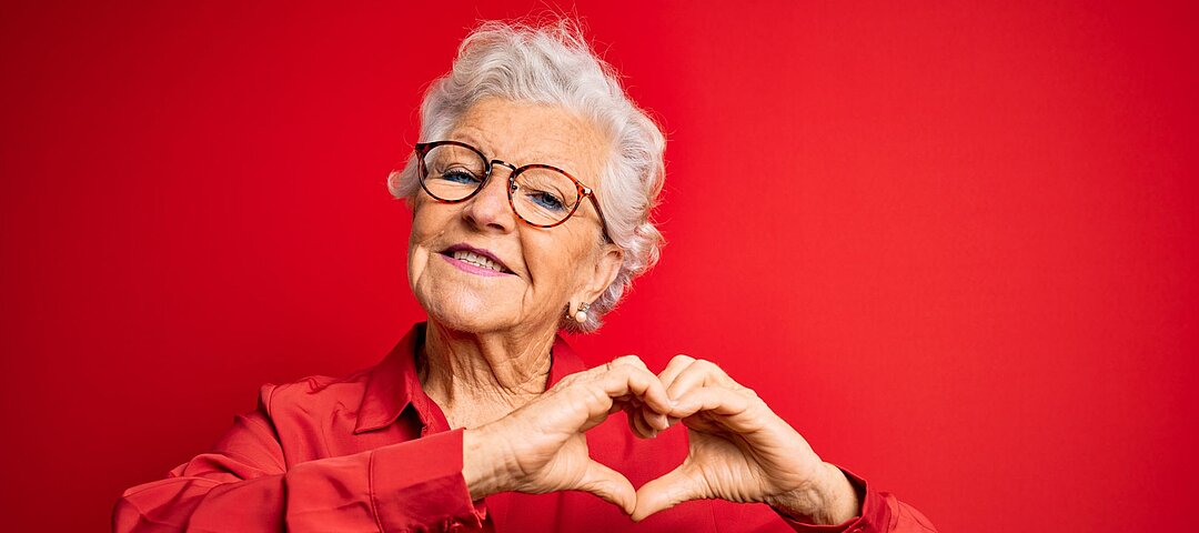 Foto: Seniorin in roter Bluse vor roter Wand macht Herzsymbol mit den Händen