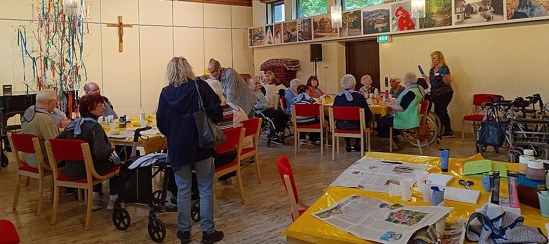 Foto: Senior:innen sitzen an mehreren Tischen mit Getränken auf roten Stühlen, im Hintergrund ein Kruzifix an der Wand