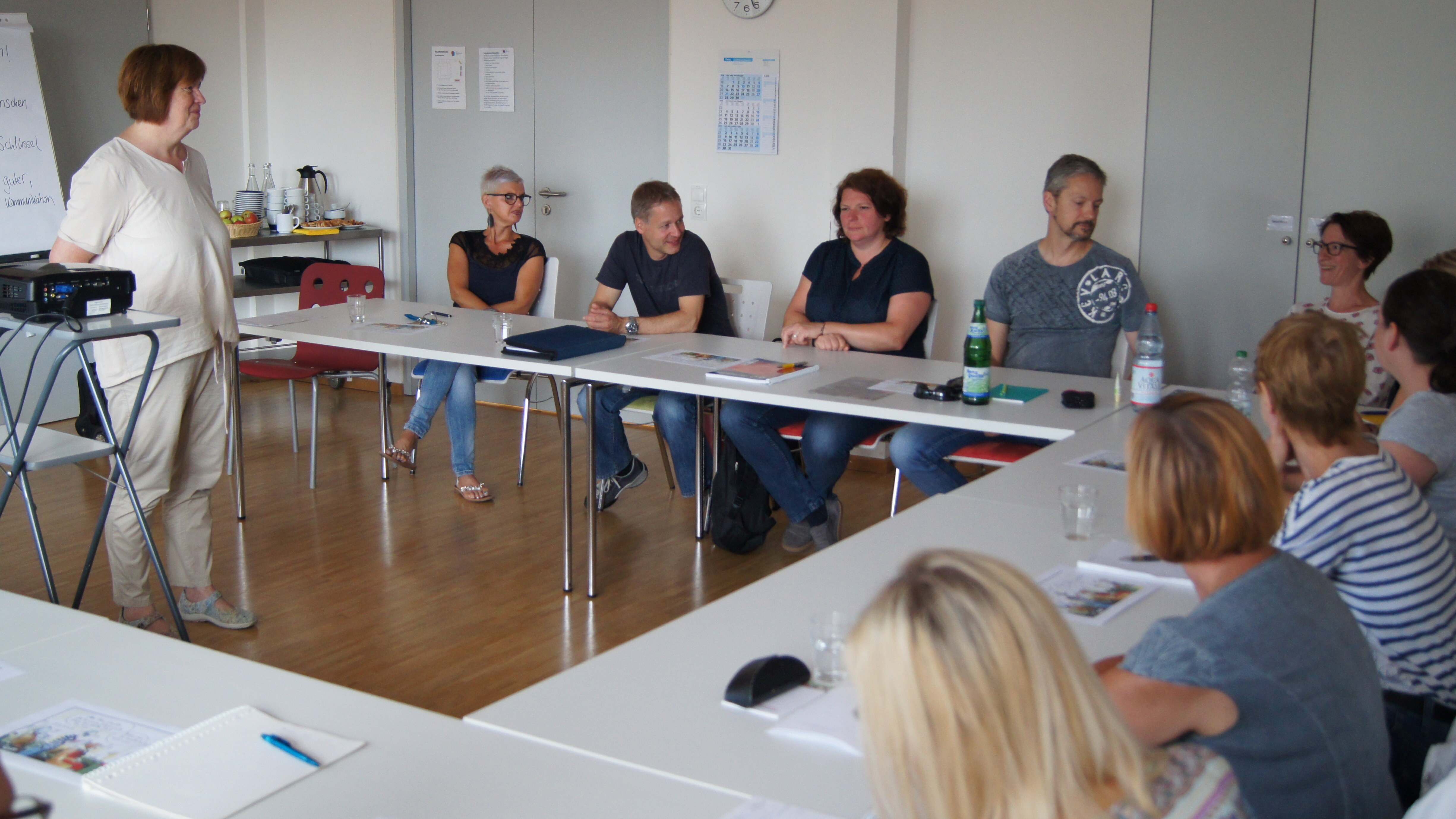 Foto: Eine Gruppe von Menschen sitzt in einer U-Form. In der Mitte steht eine Frau neben einem Beamer.