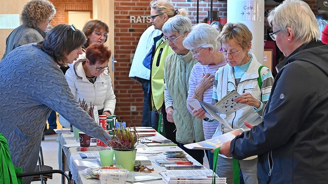 Info-Stand von PRO DEM e.V. | „Demenz-Info-Tag“, Oktober 2024 in Stuhr Foto: 9 Personen stehen um einen Tisch und schauen sich die darauf liegenden Broschüren und Flyer an.