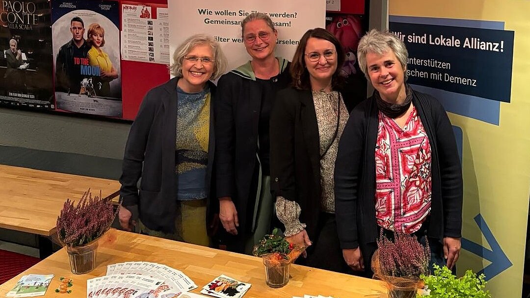 Informationsstand des Projektes MUT während der Kinovorführung „The Father“ anlässlich der Woche der Demenz 2024 (v.l.n.r: Zwei Mutmacherinnen, Projektkoordinatorin und Seniorenfachkraft der Stadt Würselen, Projektdurchführende Fachkraft) Foto: 4 Frauen stehen hinter einem Holztisch mit Broschüren und Flyern vor 2 RollUps und einer roten Stellwand