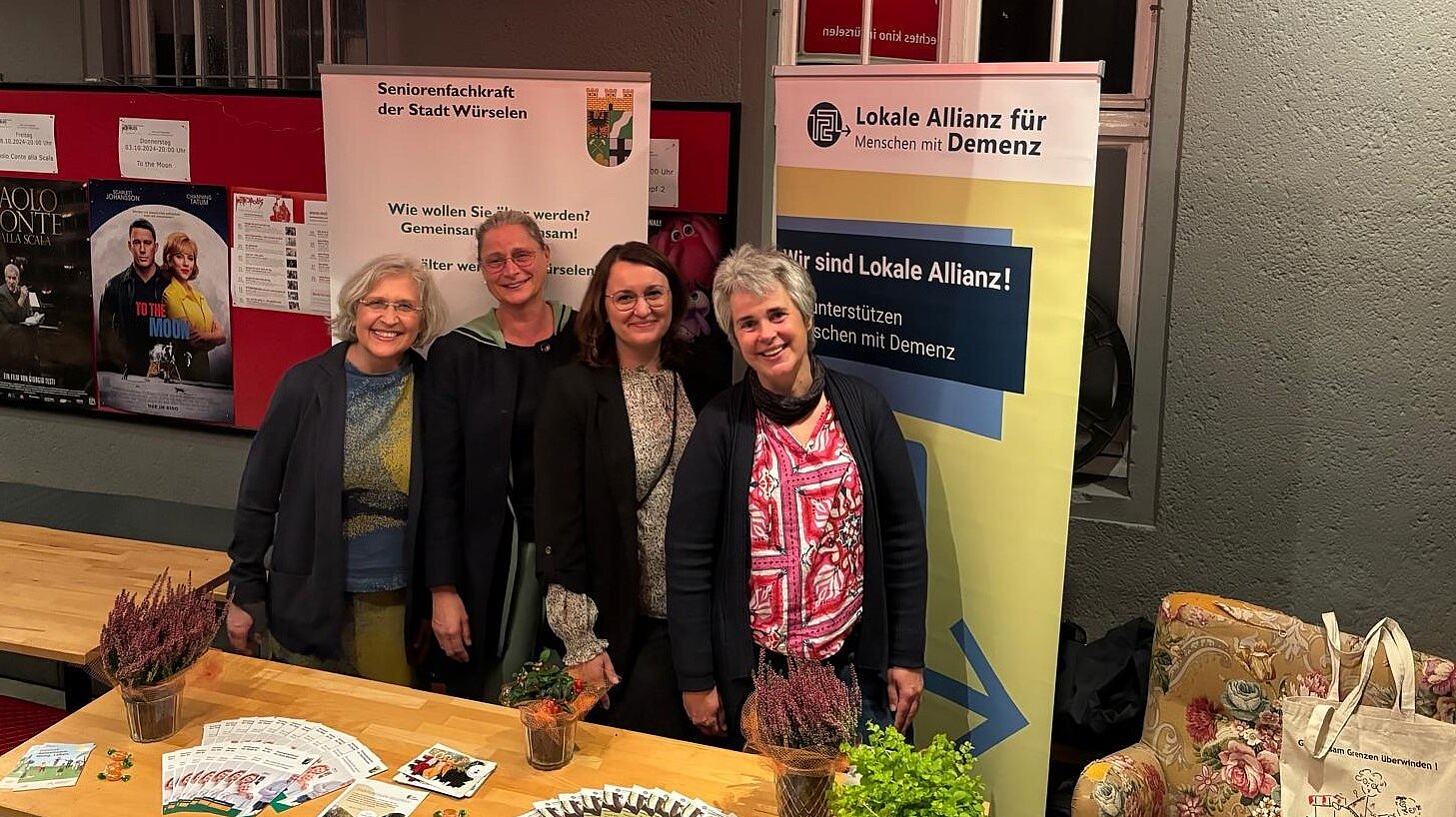 Foto: 4 Frauen stehen hinter einem Holztisch mit Broschüren und Flyern vor 2 RollUps und einer roten Stellwand