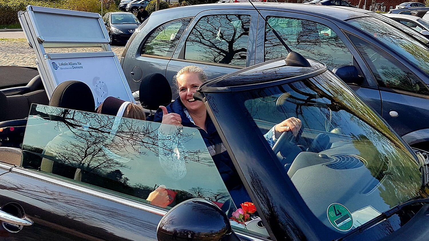 "Lokale Allianz on Tour" Foto: Parkendes Auto mit offenem Verdeck. Im Auto sitzen zwei Frauen, auf dem Rücksitz steht das Poster der Lokalen Allianz.