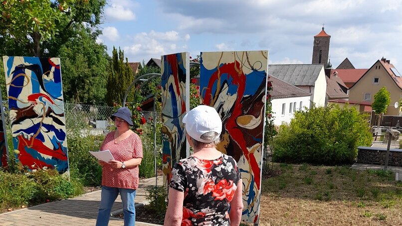 Zwei Frauen stehen vor drei großen Gemälden im Freien. Eine von ihnen hält einen Vortrag. 