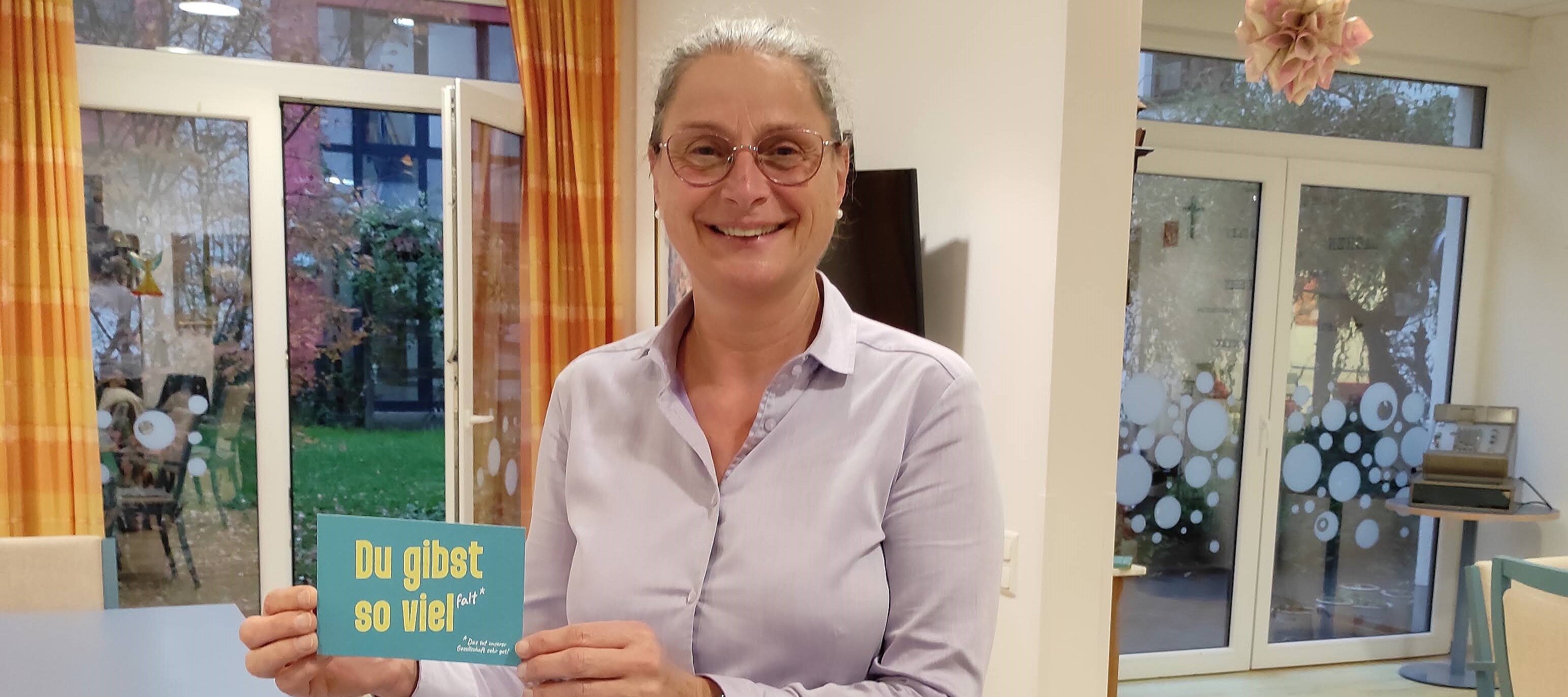 Foto: Frau steht zwischen zwei Fenstern und und hält eine Karte mit der Aufschrift „Du gibst so viel Falt“.