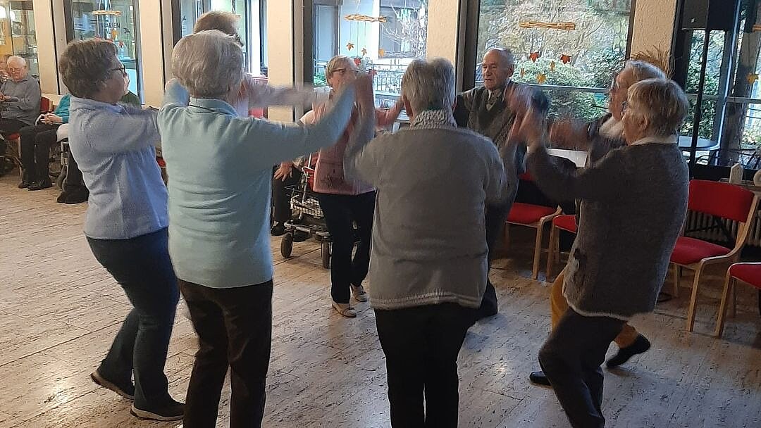 Foto: 8 Senior*innen tanzen in einem Saal im Kreis und halten sich an den Händen fest.