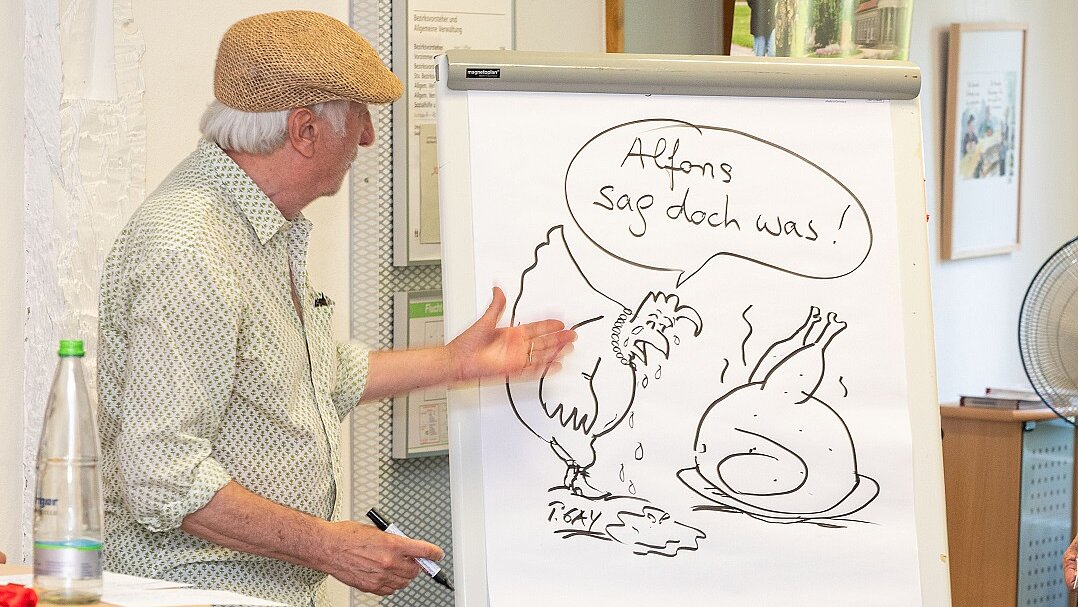 Peter Gaymann Vernissage zum 10-jährigen Jubiläum des Netzwerks Foto: Der Künstler Peter Gaymann steht vor einer Stellwand mit einer seiner Karikaturen