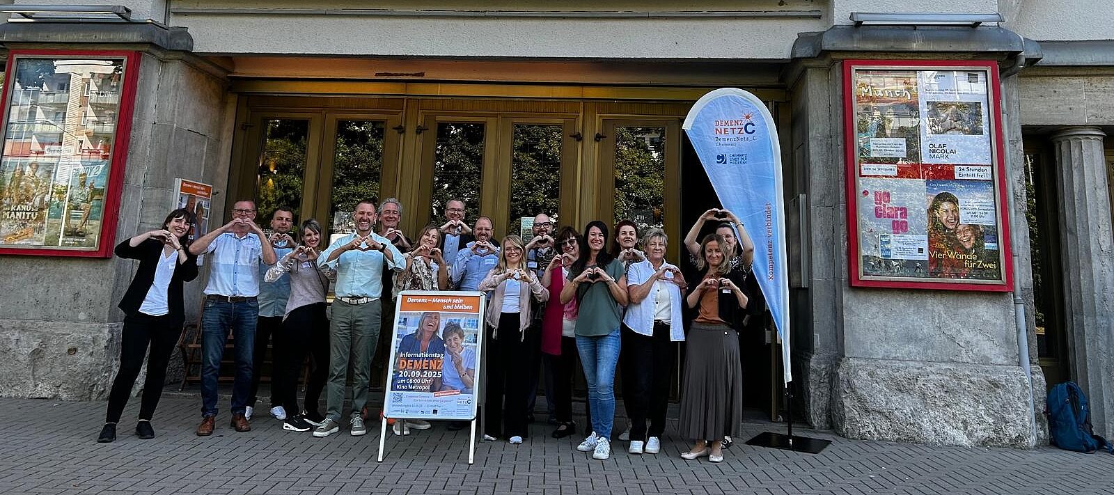 Die Netzwerkpartner stehen vor einem Kino und formen mit ihren Händen ein Herz