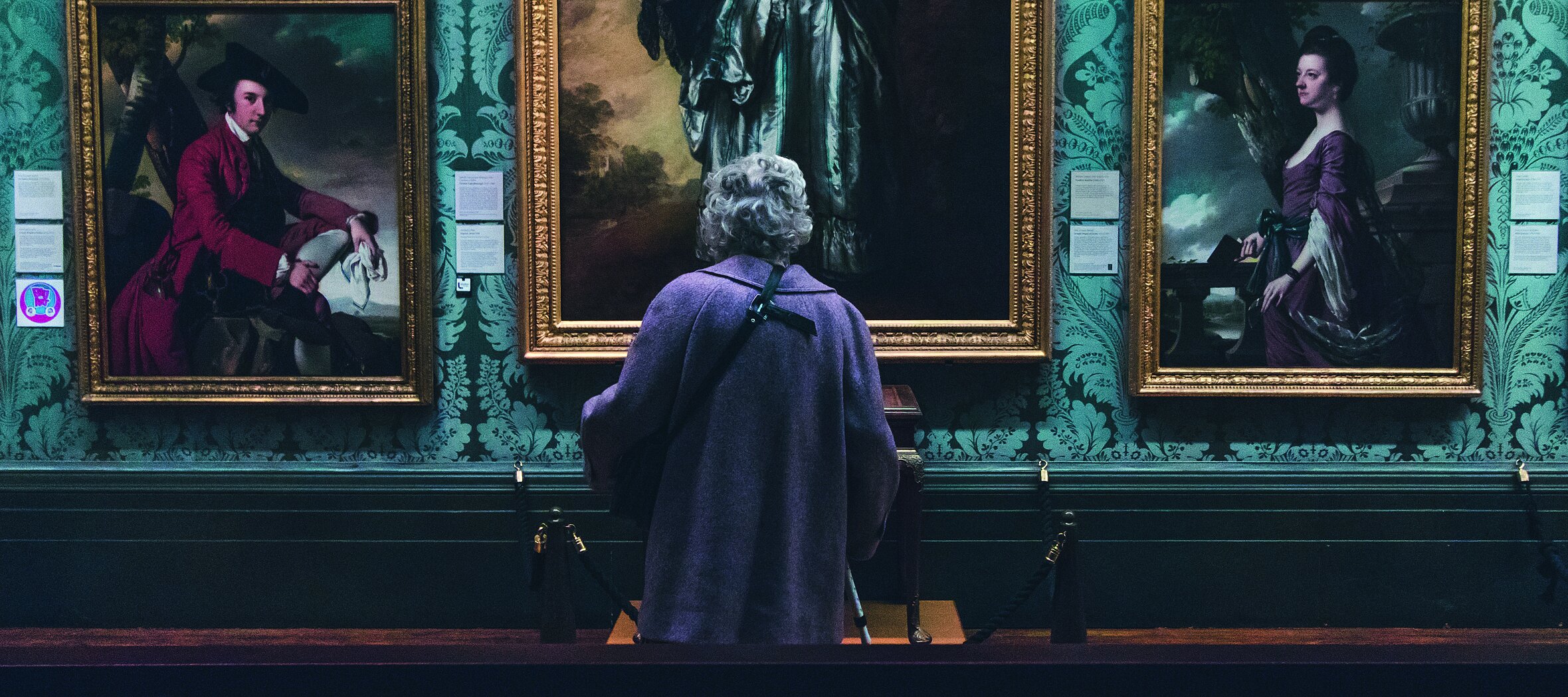 Foto: Rückenansicht einer älteren Dame, die im Museum vor einer Wand mit Ölgemälden steht 