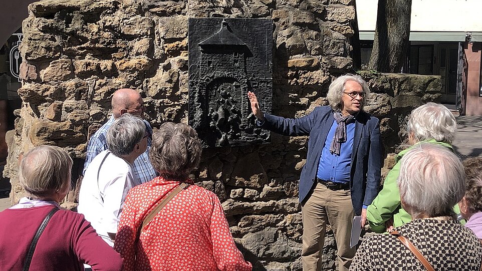 Foto: Eine Gruppe Senior:innen schauen auf eine Bronzeplakette an einer Mauer und einen mann, der dazu etwas erklärt.