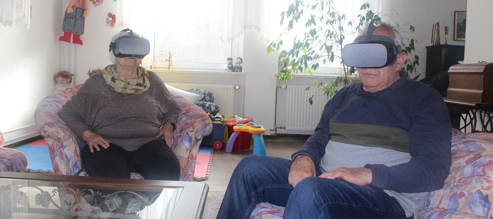 Foto: Eine ältere Frau und ein älterer Mann sitzen in einer Sitzgruppe und tragen Virtual-Reality-Brillen 