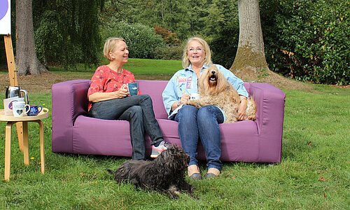 © Körber-Stiftung Auf diesem Foto sehen Sie ein lila Sofa, das draußen auf einer Wiese steht. Auf dem sofa sitzen zwei Frauen sowie zwei Hunde