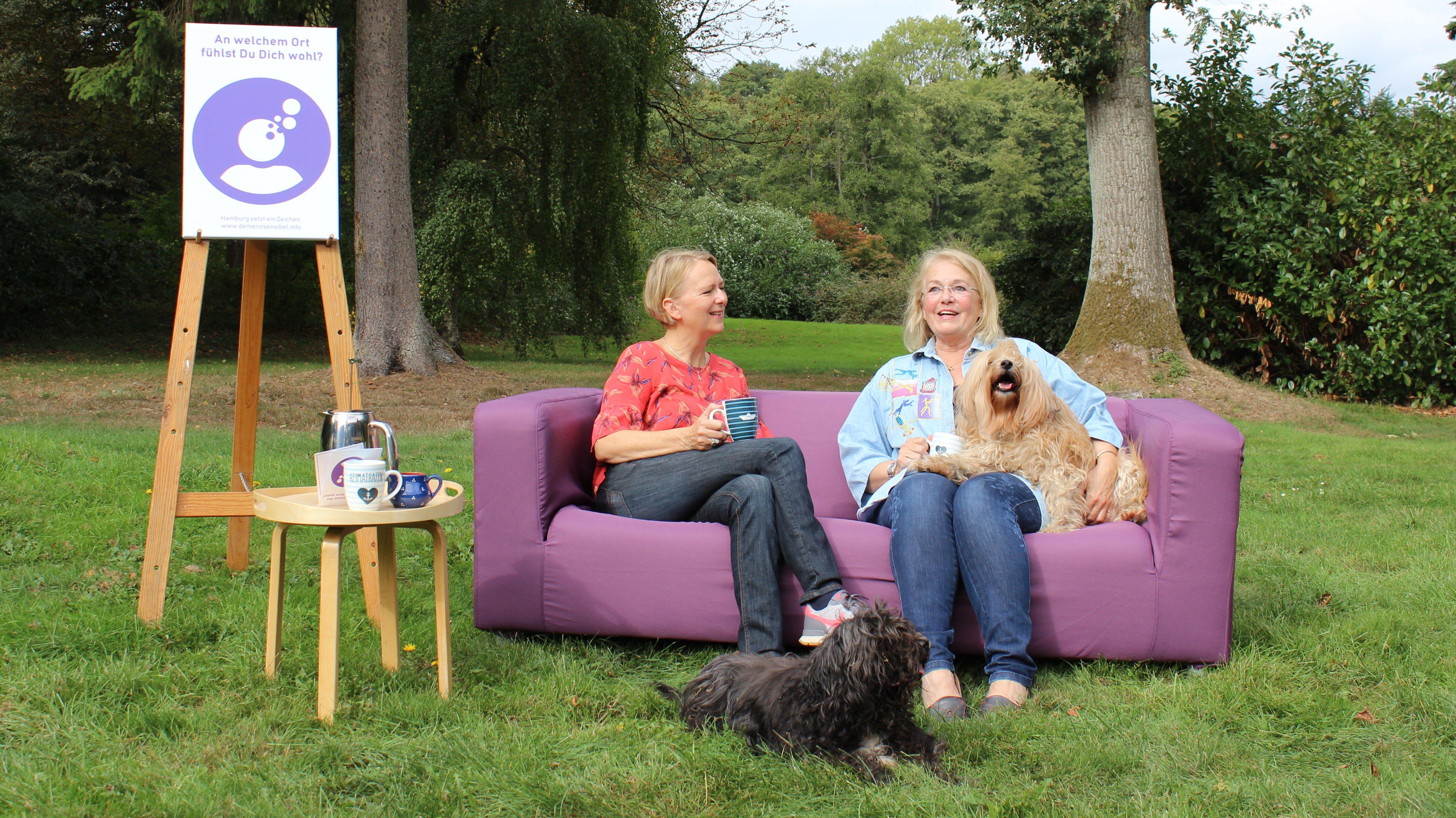 Auf diesem Foto sehen Sie ein lila Sofa, das draußen auf einer Wiese steht. Auf dem sofa sitzen zwei Frauen sowie zwei Hunde
