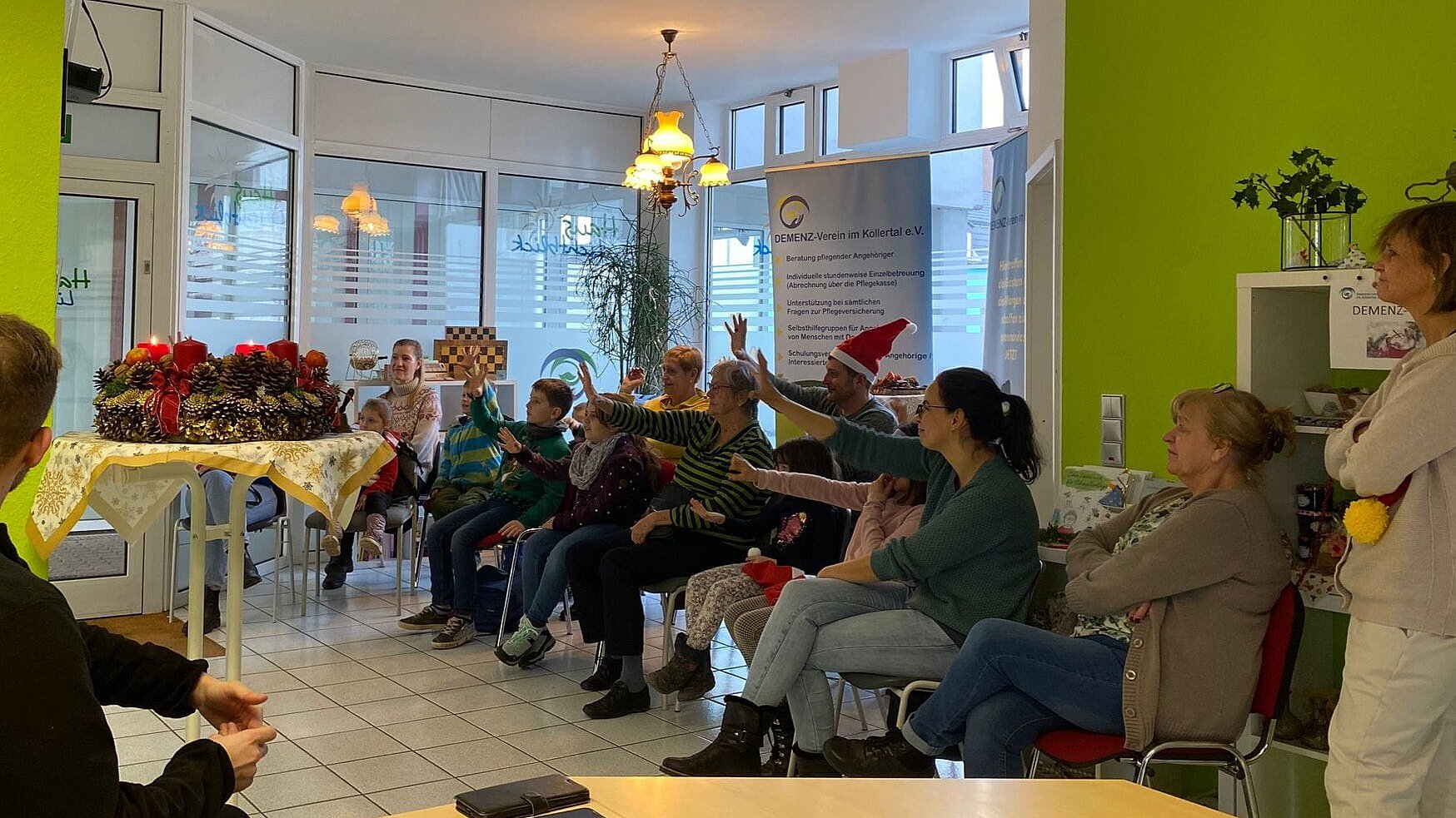 Foto: Mehrere Generationen sitzen in einem weihnachtlich geschmückten Raum vor großen Fenstern und winken einer nicht sichtbaren Person am linken Bildrand zu.