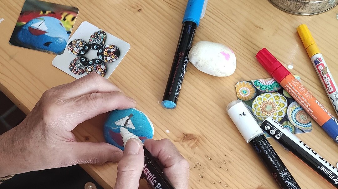 Foto: Vogelperspektive: Stifte und Steine auf einem Tisch; jemand bemalt einen Stein