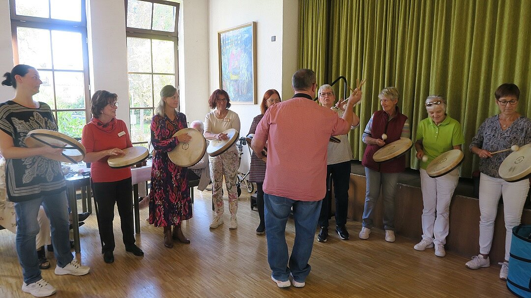 Foto: Menschen stehen in einem Raum im Kreis und schlagen auf flache Trommeln. Vor ihnen steht ein Dirigent. 