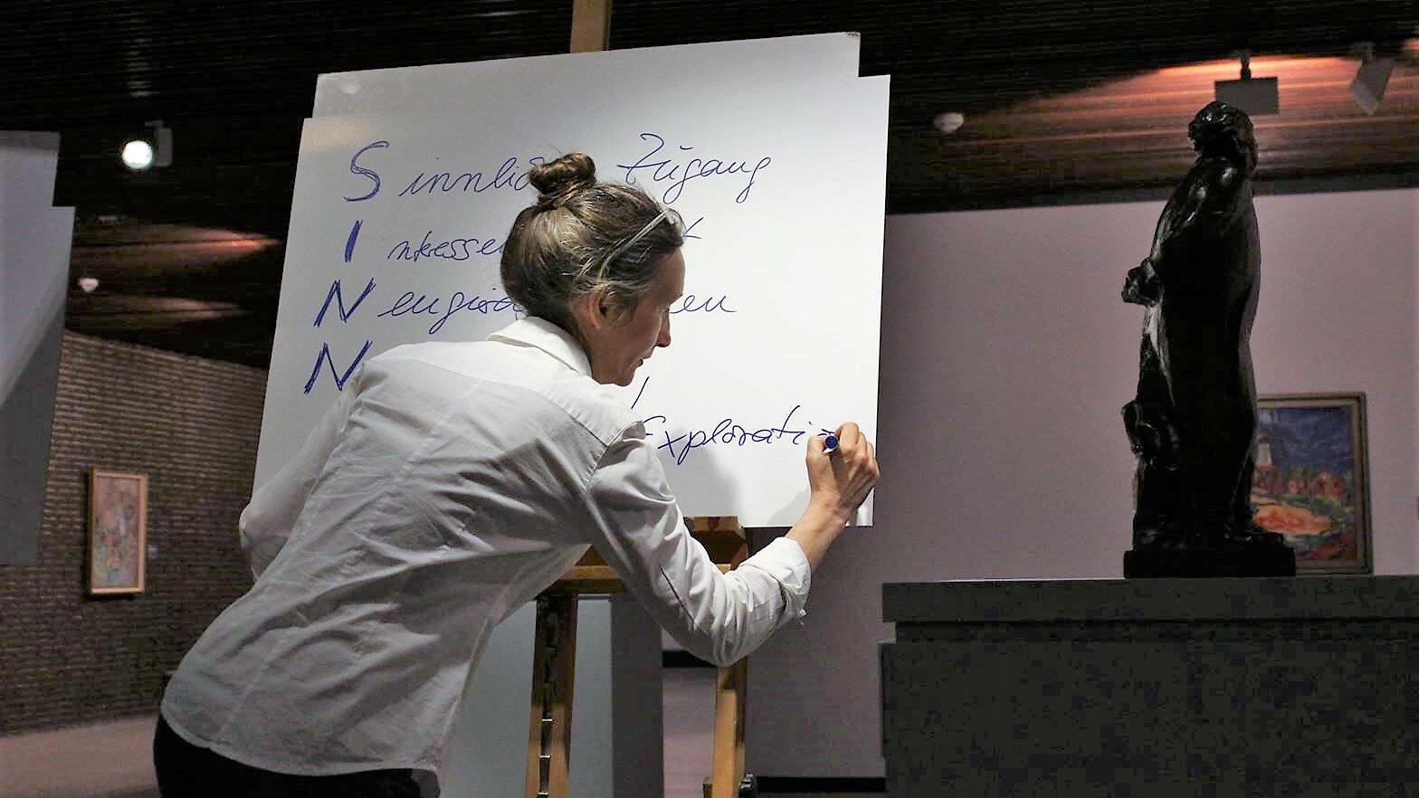 Im Vordergrund dieses Fotos schreibt eine Frau etwas auf ein Flipchart, im Hintergrund sind verschiedene Kunstwerke zu sehen