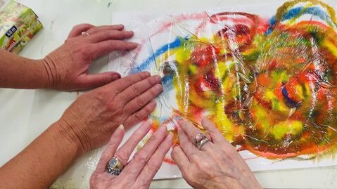 Foto: Selbst gemaltes Bild auf einem Tisch. Zwei Hände von älteren Frauen streichen eine durchsichtige Folie auf dem Bild glatt