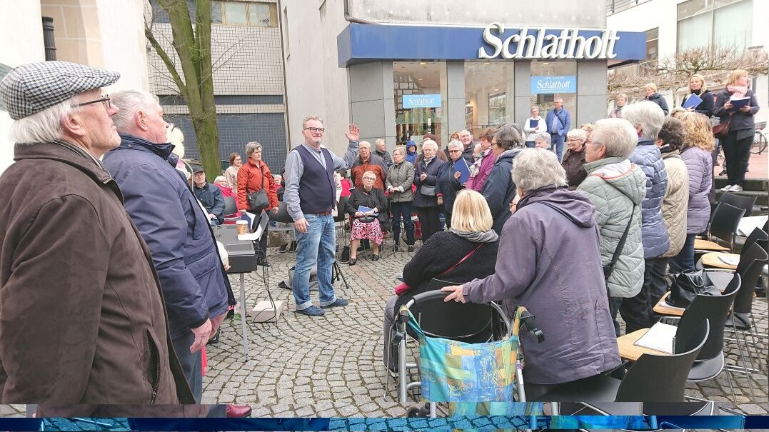 Dementiell veränderte Bürgerinnen, Angehörige und Passanten singen unter der Leitung von Jürgen Kleinschmidt im Frühjahr 2019 vor der Stadtkirche St. Georg in Lünen Auf diesem Foto sehen Sie einen öffentlichen Platz, auf dem viele ältere Menschen gemeinsam singen