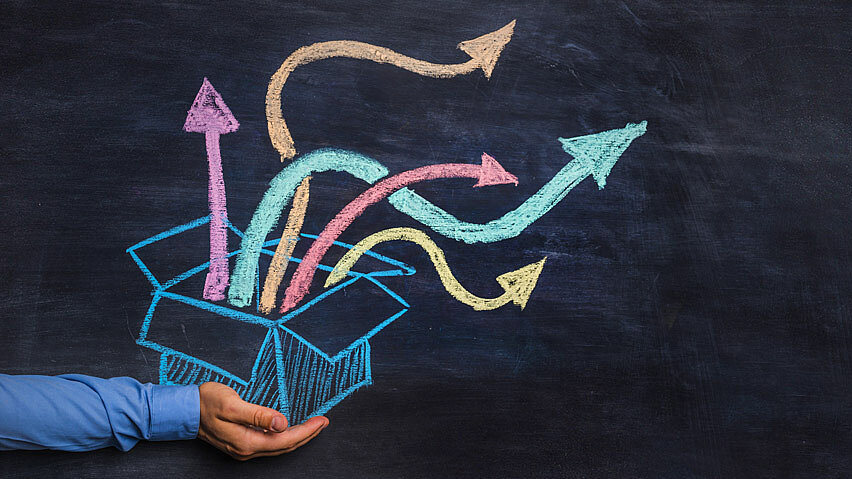 © istock.com_warchi Schwarze Schultafel: ein mit bunter Kreide gemalter geöffneter Karton, aus dem bunte Pfeile herauskommen. Zu sehen ist weiterhin die Hand einer realen Person, die scheinbar den Karton in den Händen hält