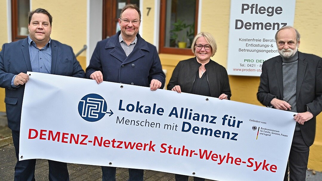 Netzwerkgründung in Weyhe, 2022 (v.l.n.r.): Weyhes Bürgermeister Frank Seidel, seine Kolleg*innen Stephan Korte aus Stuhr und Suse Laue aus Syke sowie PRO DEM-Vorsitzender Frithjof Troue Foto: 4 Personen stehen vor einem gelben Haus und halten ein großes Banner "DEMENZ-Netzwerk Stuhr-Weyhe-Syke"