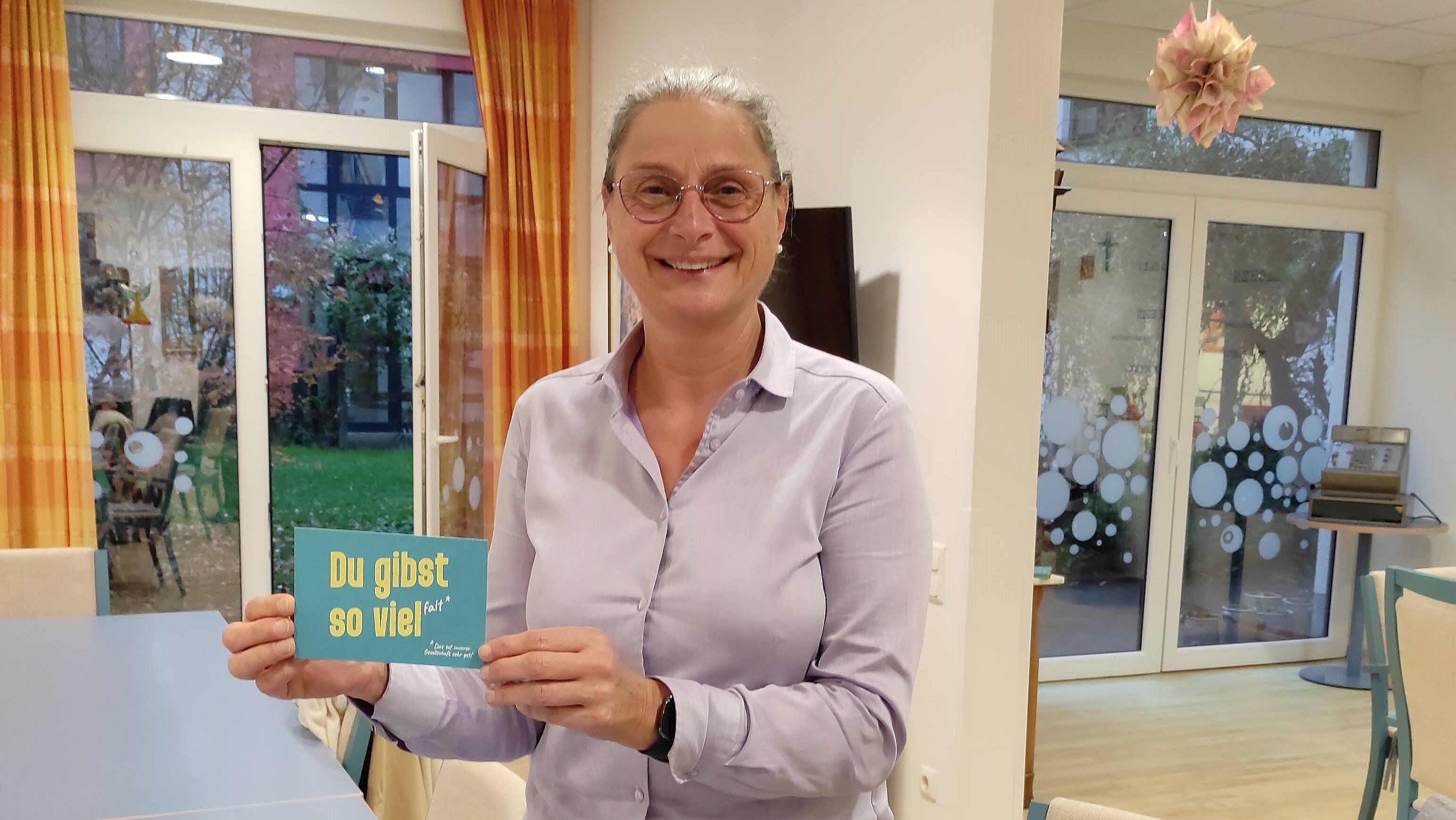 Foto: Frau steht zwischen zwei Fenstern und und hält eine Karte mit der Aufschrift „Du gibst so viel Falt“.