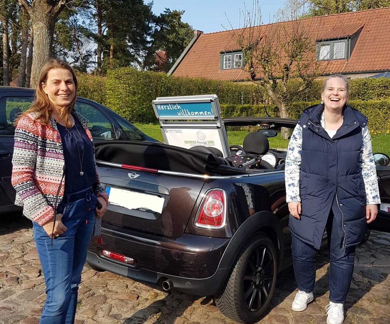 Parkendes Auto mit offenem Verdeck und Poster der Lokalen Allianz auf dem Rücksitz. Rechts und links stehen zwei Frauen und lachen in die Kamera.