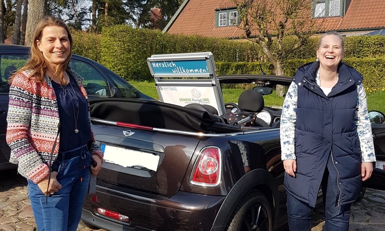 Parkendes Auto mit offenem Verdeck und Poster der Lokalen Allianz auf dem Rücksitz. Rechts und links stehen zwei Frauen und lachen in die Kamera.