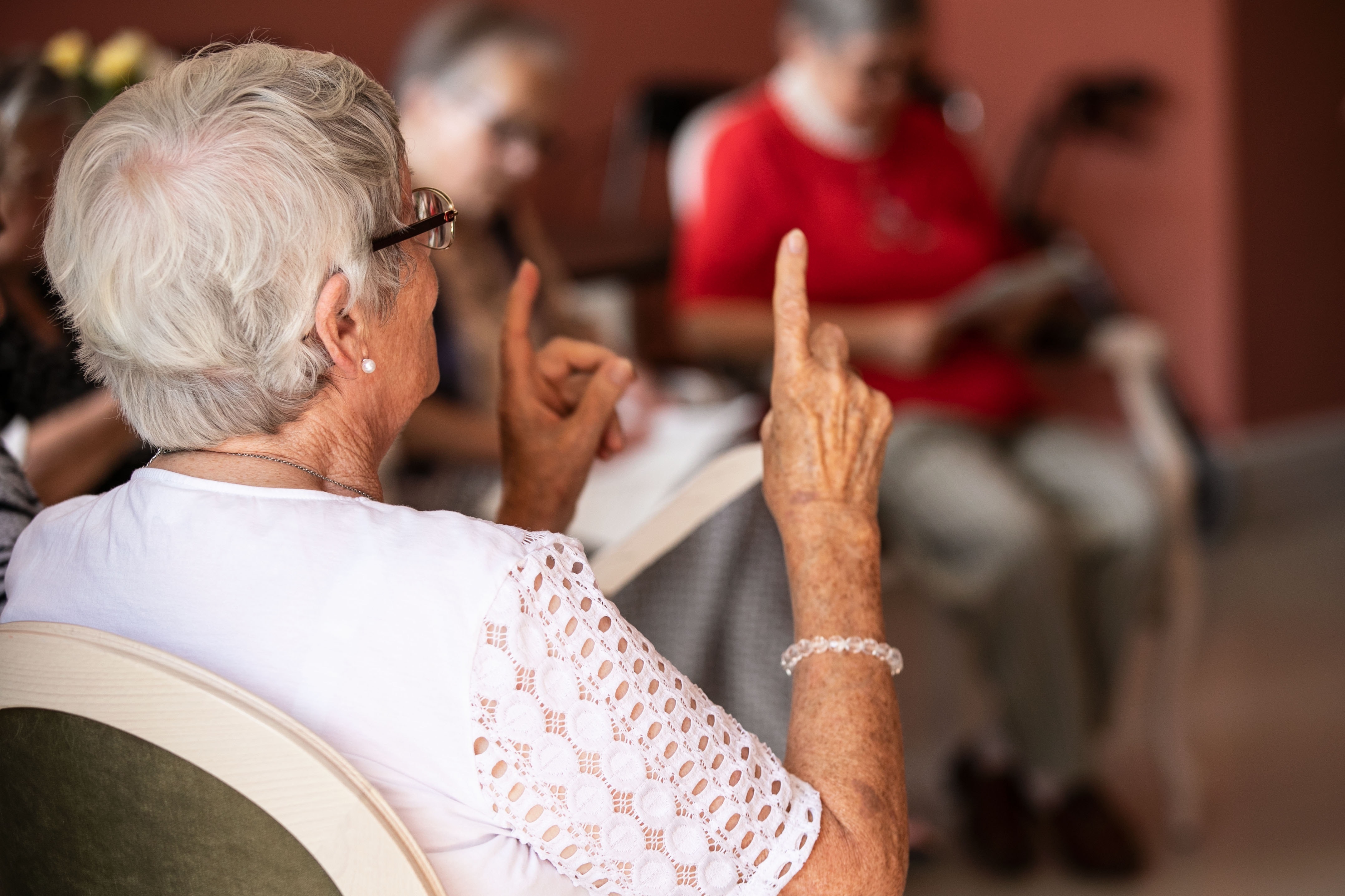 Foto: Ältere sitzende Frau von hinten, die ihre Zeigefinger in die Luft hält 