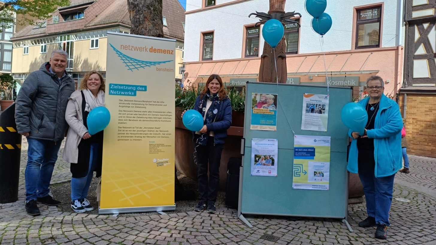 Foto: Vier Personen stehen mit Roll-Up und Stellwand auf einem öffentlichen Platz. 