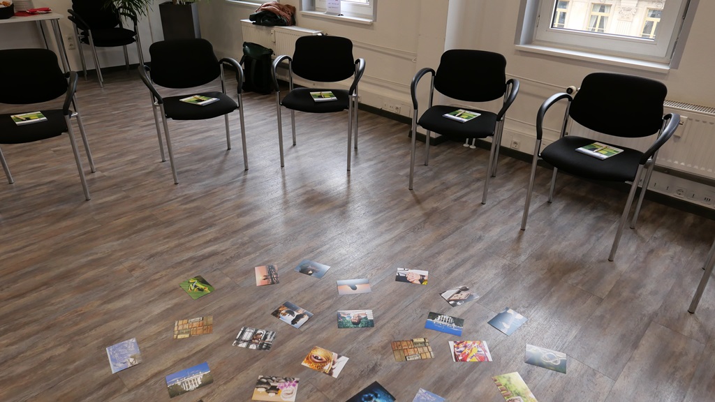 Leere Stühle im Kreis. Auf dem Boden in der Mitte liegen verschiedene Postkarten