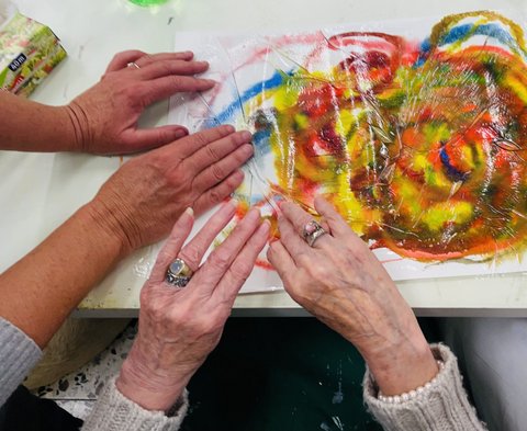 Selbst gemaltes Bild auf einem Tisch. Zwei Hände von älteren Frauen streichen eine durchsichtige Folie auf dem Bild glatt